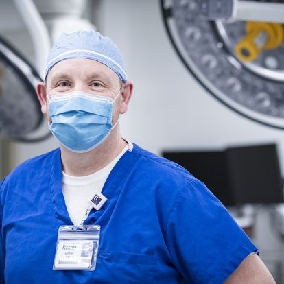 Ronnie Lott is pictured standing inside one of the operating rooms at the KGH site. He has blue eyes and is wearing blue scrubs, a scrub hat and mask.