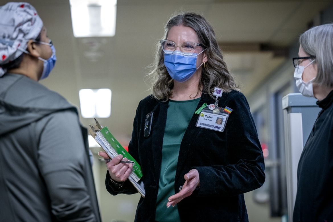 Robin Westphal is pictured standing in the middle of two colleagues in the Intensive Care Unit. Robin has dirty blonde hair with grey streaks, blue eyes and wears glasses. She’s wearing a green blouse with a black blazer on top. All three individuals are wearing masks.