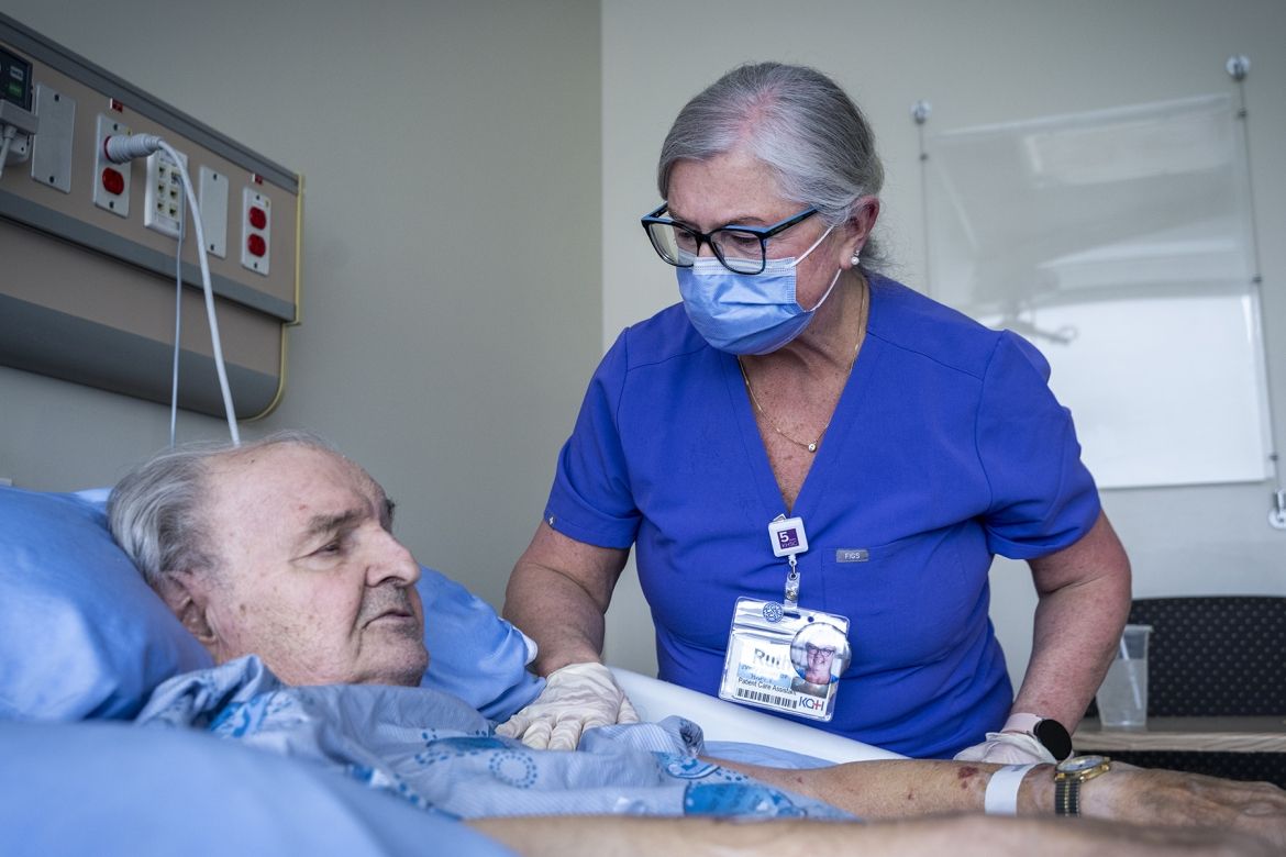 Ruth Bradshaw is pictured caring for a patient who is lying in a hospital bed. Bradshaw has white/silver hair, pulled back into a bun, black framed glasses and is wearing blue scrubs with a mask. She’s leaning over the patient in the bed, and her hand is on his upper arm in a loving way.