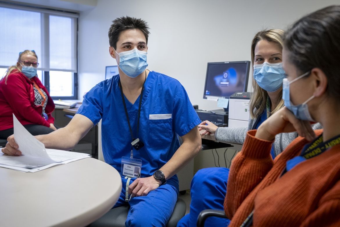 Evan Earl is seated next to two women around a circular table, another woman is seen seating in the background. Earl has short, dark, spiky hair, brown eyes, and is wearing a mask and bright blue scrubs. The two woman are also wearing masks and bright blue scrubs.