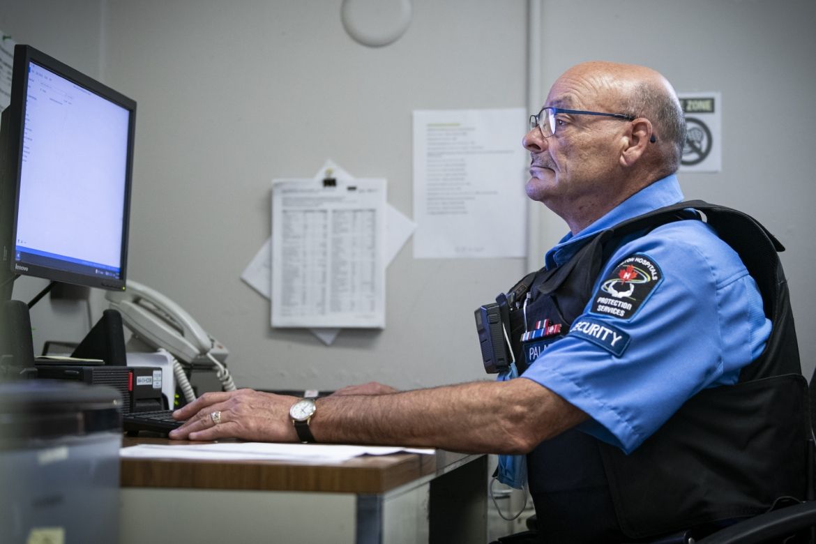 Mario Lelievre is pictured sitting at his desk looking at a monitor at Hotel Dieu Hospital. He has brown eyes, wears blue glasses and is bald. He’s wearing his Paladin Security uniform, which includes a blue collared, button down shirt, protective vest, radio, and ID badge.