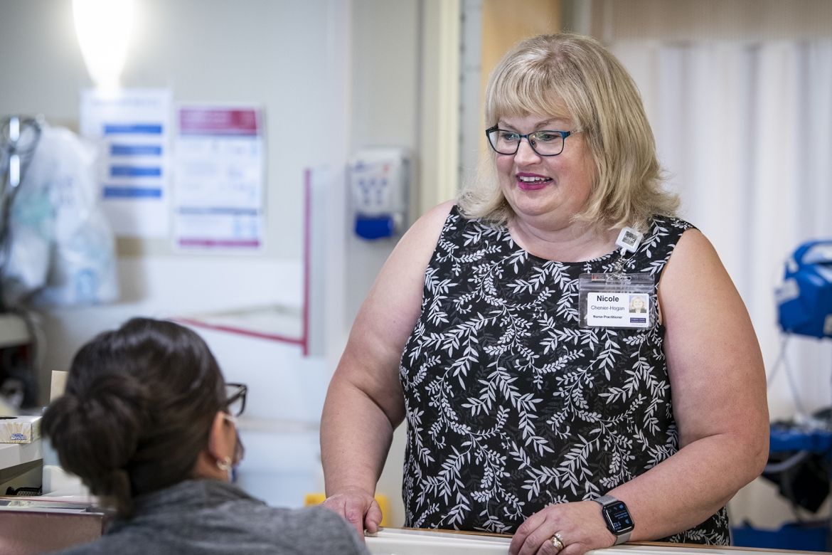Nicole Chenier-Hogan is pictured standing next to a unit care desk. She’s looking down and speaking with a nurse sitting at the care desk.