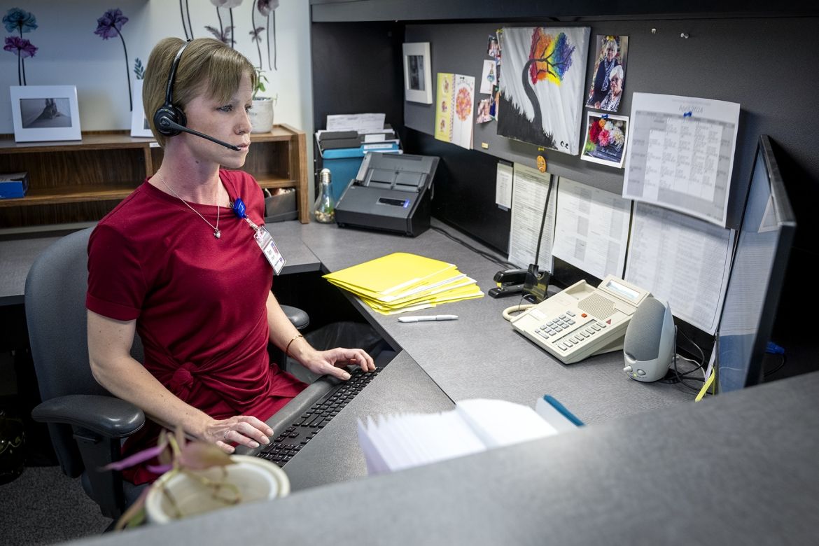 Taryn Oegema is pictured sitting at her desk, looking at her computer. She has a headset on and her fingers are typing on her keyboard. She has short blonde hair, blue eyes and is wearing a dark red dress.