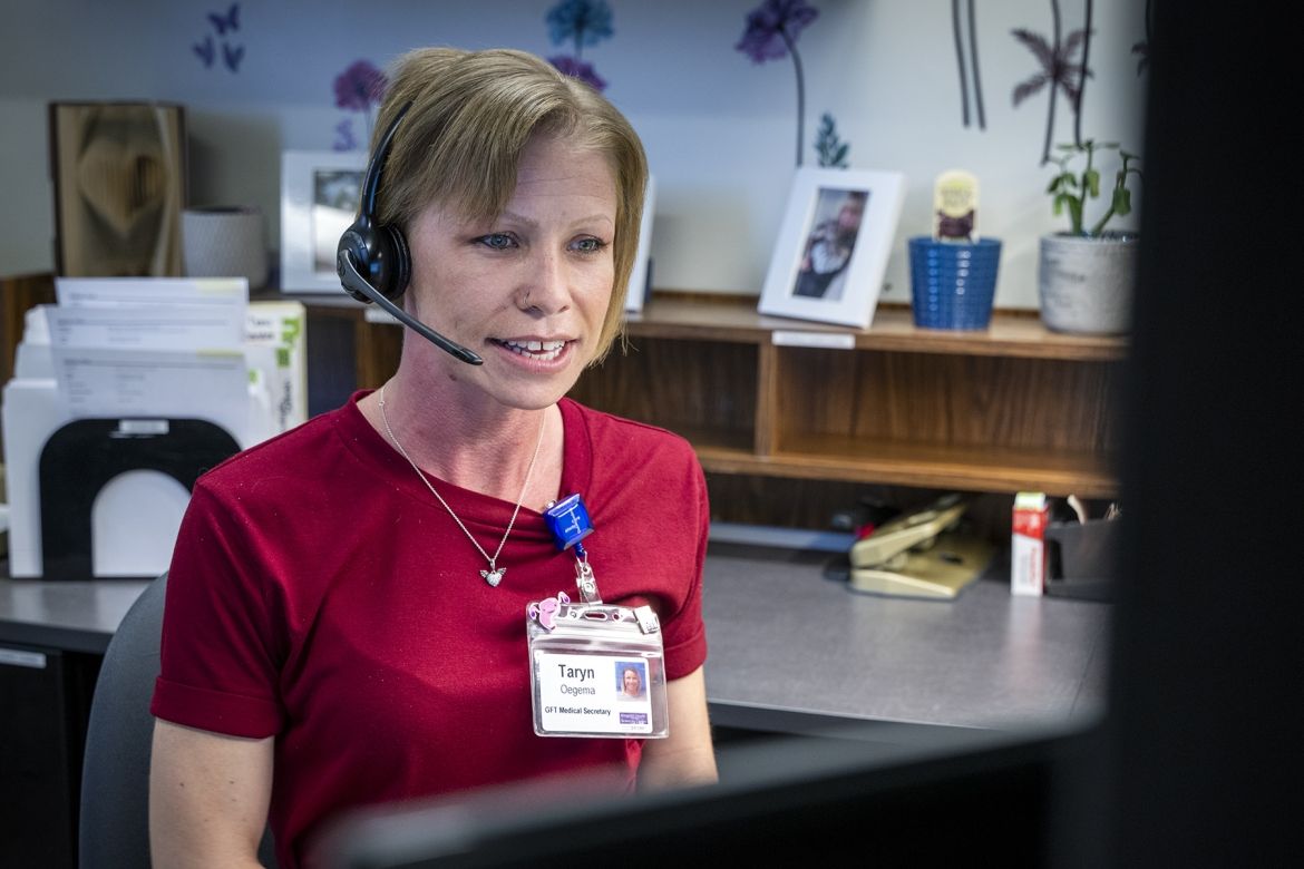 Taryn Oegema is pictured sitting at her desk, looking at her computer. She has a headset on and is talking on the phone. She has short blonde hair, blue eyes and is wearing a dark red dress.