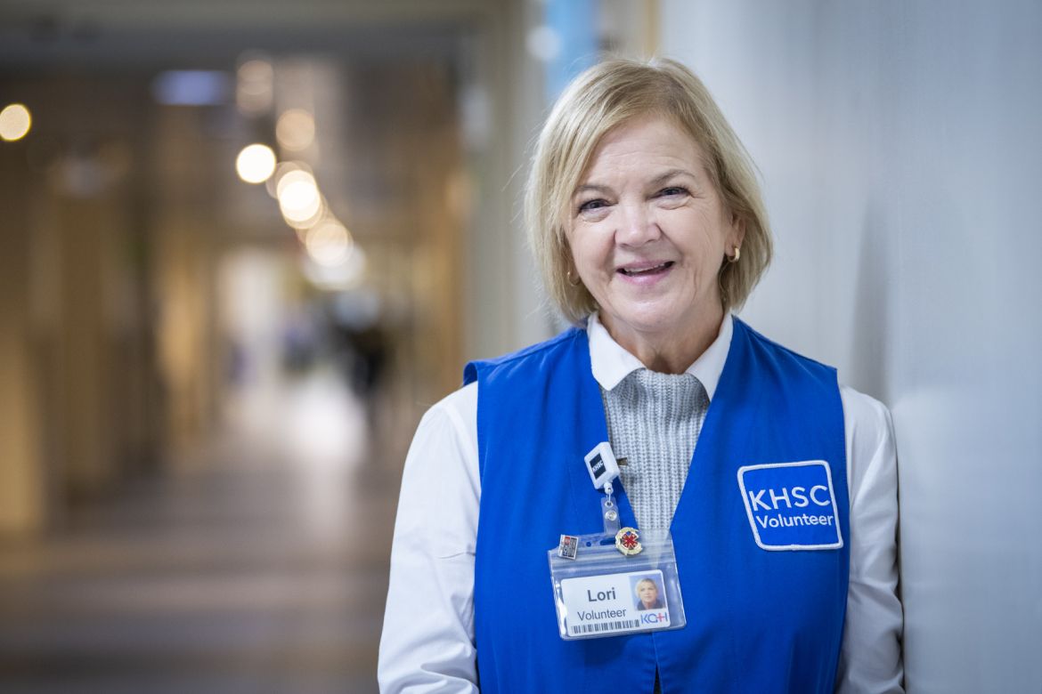 Hospital volunteer wearing a blue Kingston Health Sciences Centre volunteer vest and name badge, standing in a hospital hallway.