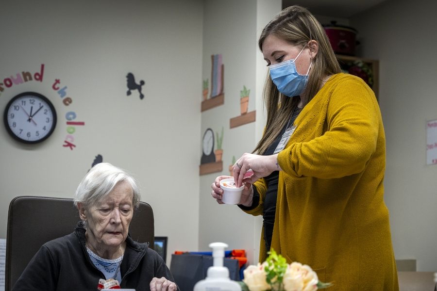 Jessica McCutcheon is standing in front of a table opening art supplies for an elderly female patient, who is seated. McCutcheon has dark blonde hair and brown eyes. She’s wearing a black top, mustard cardigan and a mask over her mouth and nose.