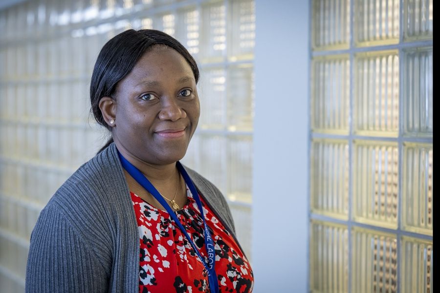 Christiana Adewale is pictured standing in a hallway at the Kingston General Hospital Site. She’s Black, has long-dark hair that’s tied up in a low ponytail and dark brown eyes. She’s wearing a bright red, white and black patterned blouse with a grey cardigan on top.