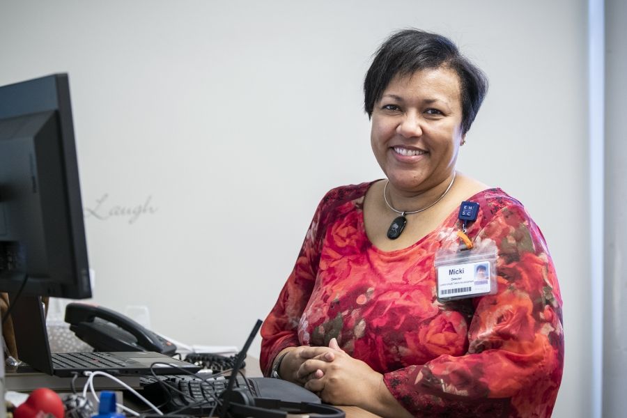 Micki Mulima is pictured sitting at her desk at the Kingston General Hospital site. She’s Black, has short-dark hair and brown eyes. She’s wearing a bright red, patterned blouse with roses.