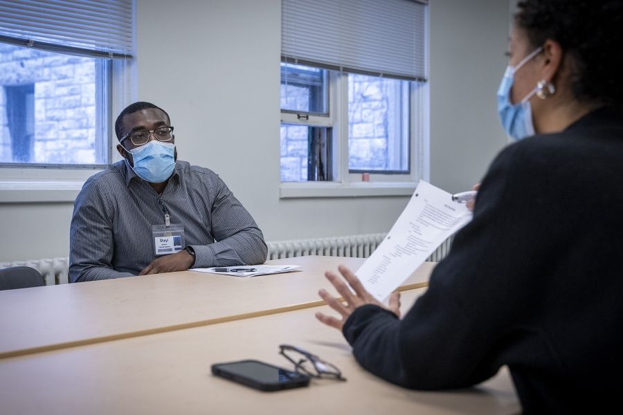 Members of the Black Staff Community Group are seated around a boardroom table as they meet. The image shows a Black man in focus sitting directly across from a Black woman who is holding up a piece of paper for him to see.
