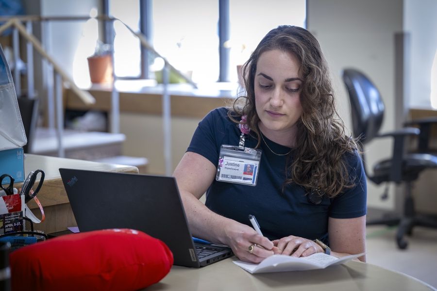 Justine White is seated at a table with her computer, a cardiac surgery pillow and some paper, which she’s jotting down notes on. She has shoulder length, medium brown, wavy hair and brown eyes. She’s wearing a navy blue shirt with her KHSC ID badge.