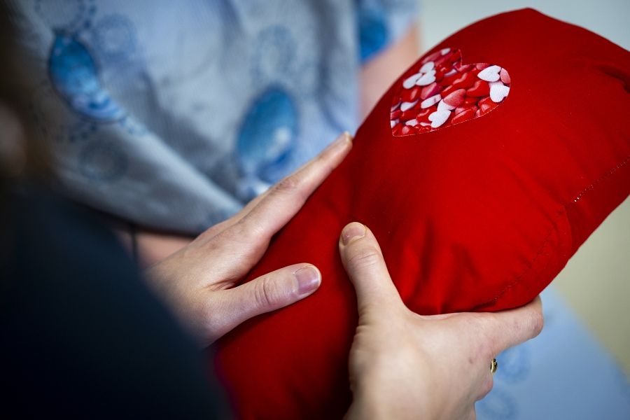 A close up shot of Justine White’s hands holding a cardiac surgery pillow. The pillow is bright red with a heart near the top of the pillow.