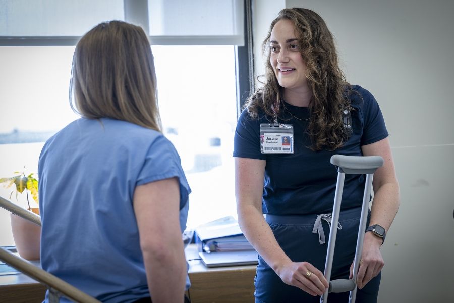 Justine White is standing and putting together a crutch, while looking at physiotherapy assistant Danielle Knapton, whose back is to the camera. White has shoulder length, medium brown, wavy hair and brown eyes. She’s wearing a navy blue shirt with her KHSC ID badge.