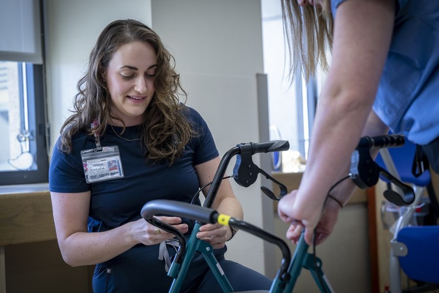 Justine White is kneeling working on a walker, a mobility aid device. Physiotherapy assistant Danielle Knapton, whose back is to the camera, is holding the device. White has shoulder length, medium brown, wavy hair and brown eyes. She’s wearing a navy blue shirt with her KHSC ID badge.