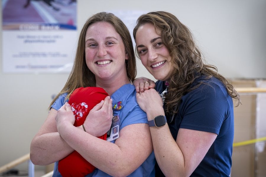 Justine White and physiotherapy assistant Danielle Knapton embracing with a cardiac surgery pillow. White has shoulder length, medium brown, wavy hair and brown eyes. She’s wearing a navy blue shirt with her KHSC ID badge. Knapton has straight medium brown hair and blue eyes. She’s wearing light blue scrubs with her KHSC ID badge.