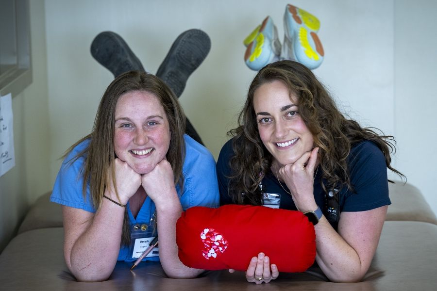 Justine White and physiotherapy assistant Danielle Knapton are laying side-by-side with their feet crossed in the air, holding a cardiac surgery pillow. White has shoulder length, medium brown, wavy hair and brown eyes. She’s wearing a navy blue shirt with her KHSC ID badge. Knapton has straight medium brown hair and blue eyes. She’s wearing light blue scrubs with her KHSC ID badge.