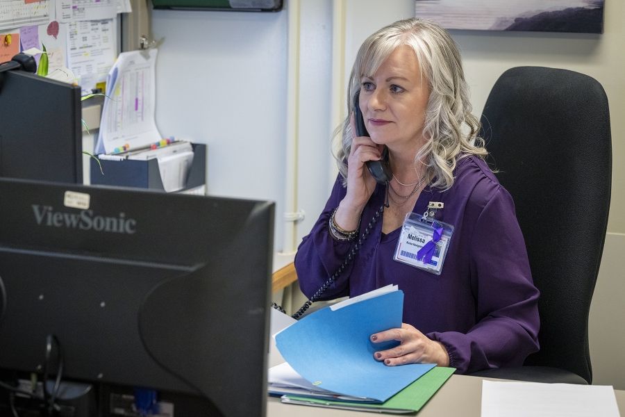 Melissa Meisner is sitting behind a desk, making a phone call. She’s holding the receiver near her ear with one hand, while the other is going through a folder. Meisner has blue eyes, shoulder length, silver hair and is wearing a bright purple blouse.