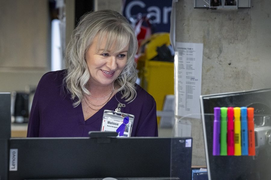 Melissa Meisner is smiling standing behind a care desk. She’s looking down at a monitor. Meisner has blue eyes, shoulder length, silver hair and is wearing a bright purple blouse.