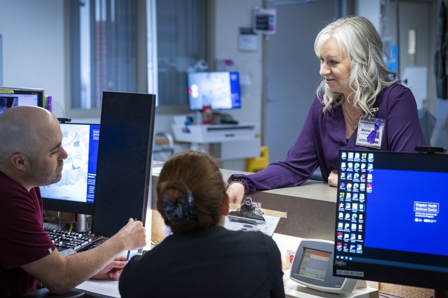 Melissa Meisner is chatting with two colleagues who are sitting at a care desk. Meisner is standing behind the desk, opposite from them. Meisner has blue eyes, shoulder length, silver hair and is wearing a bright purple blouse.