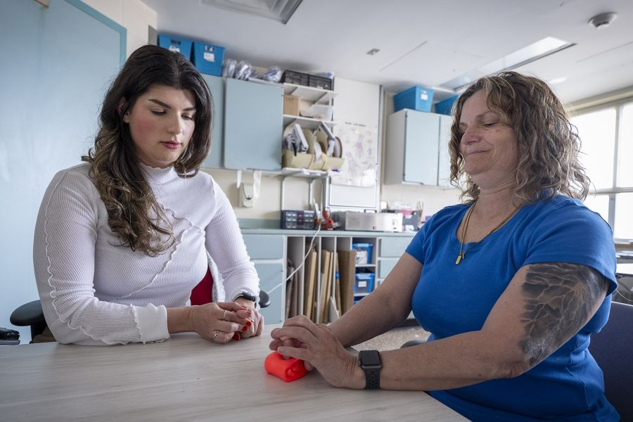 Andreea Bocicariu is seated on a table, next to a patient in the Orthopedics and Plastics Clinic. The patient is doing a hand exercise with a bright orange tool. Bocicariu is observing her patient and looking directly at her hands.