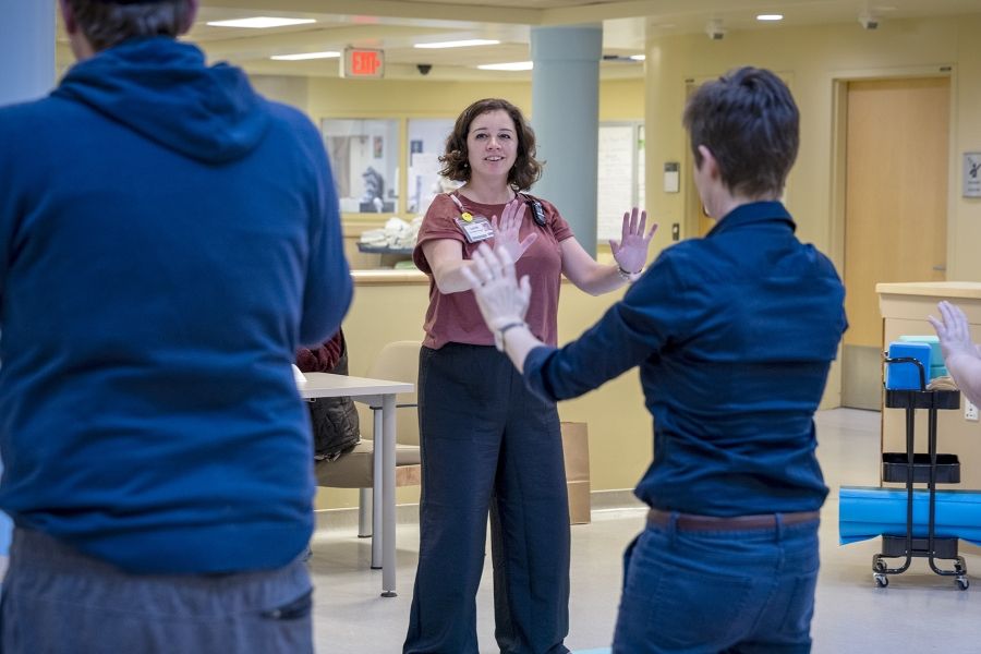 Laurie Hill is photographed leading her mindful movement group. Hill is standing with her arms out. She has short brown wavy hair and is wearing a dusty rose blouse and black pants.
