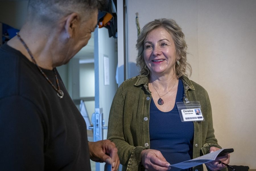 Christine Hennigar is smiling, talking to a guest at Geaganano Residence. She has light blonde, wavy hair, blue eyes and is wearing a navy blue top with an army green jacket. She’s holding a clipboard.