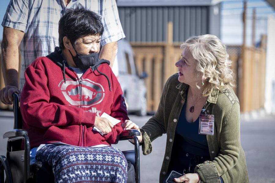 Christine Hennigar is crouched down, speaking with a guest in a wheelchair. They’re photographed outside of Geaganano Residence.