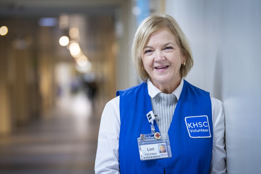 Hospital volunteer wearing a blue Kingston Health Sciences Centre volunteer vest and name badge, standing in a hospital hallway.