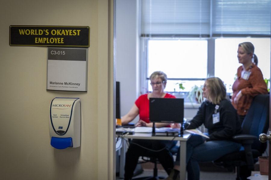 A wide shot outside Marianne McKinney’s office. In focus is a shot of a sign that says “World’s Okayest Employee”, in the background out of focus is McKinney with two other colleagues, sitting at her desk.
