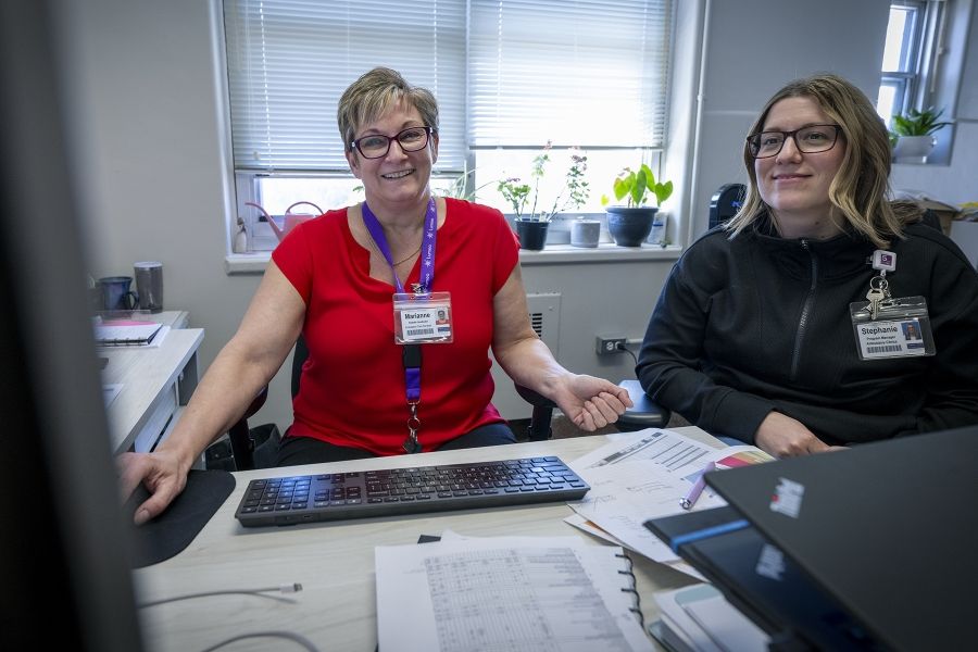 A shot of Marianne McKinney sitting at her desk with a colleague. McKinney is smiling and looking at computer monitor. McKinney has short dirty blonde hair, blue eyes, wears glasses and is dressed in a bright red blouse. Her colleague is smiling looking at the same computer monitor.