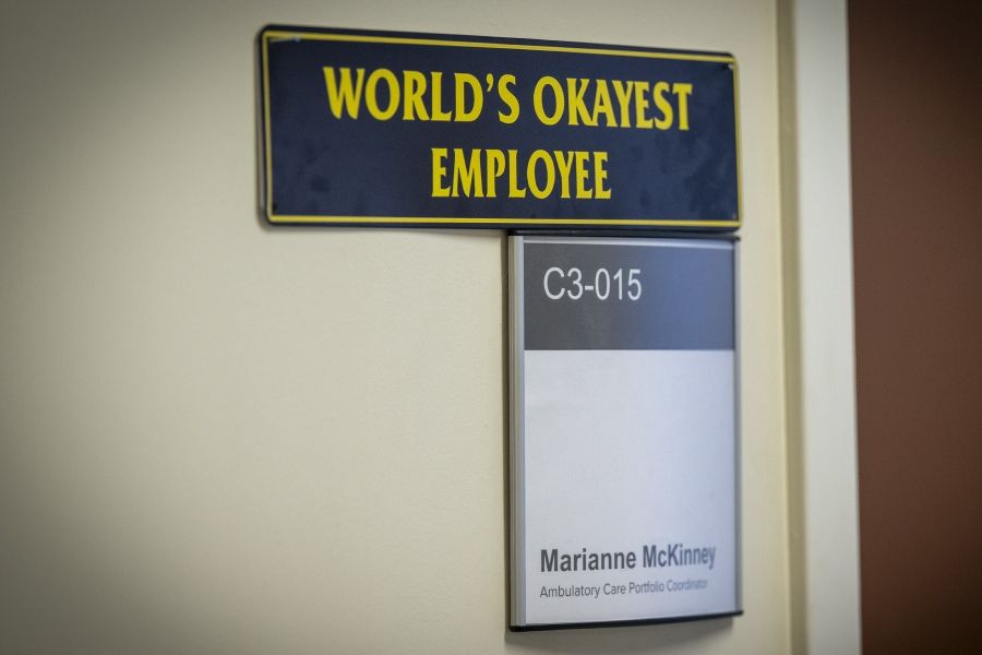 A close up shot of a sign outside of Marianne McKinney’s office that says “World’s Okayest Employee.” The sign is black with bright yellow lettering.