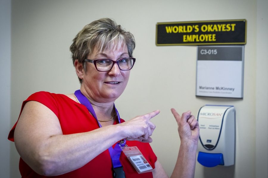 A shot of Marianne McKinney standing outside her office pointing to a sign that says “World’s Okayest Employee.” McKinney has short dirty blonde hair, blue eyes, wears glasses and is dressed in a bright red blouse.