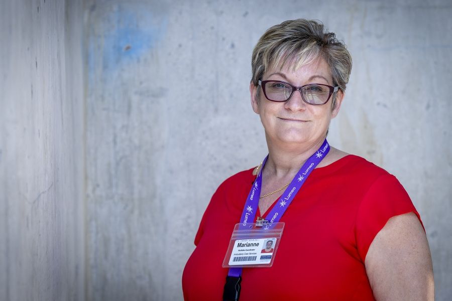A mid shot of Marianne McKinney standing in a hallway, smiling directly at the camera. McKinney has short dirty blonde hair, blue eyes, wears glasses and is dressed in a bright red blouse.