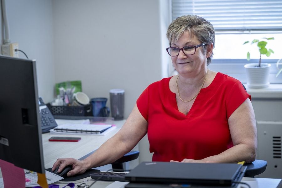 A shot of Marianne McKinney sitting at her desk, smiling at her computer monitor. McKinney has short dirty blonde hair, blue eyes, wears glasses and is dressed in a bright red blouse.