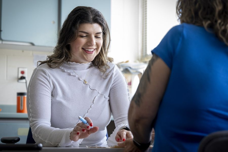 Andreea Bocicariu is seated on a table across from her patient, whose back is to the camera, in the Orthopedics and Plastics Clinic. Bocicariu is smiling, while looking down at some paperwork with a pen in her hand.