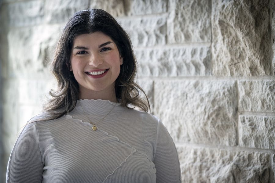 Andreea Bocicariu is standing against a wall and smiling directly into the camera. She has brown wavy hair, brown eyes and is wearing a white blouse with a gold heart necklace.