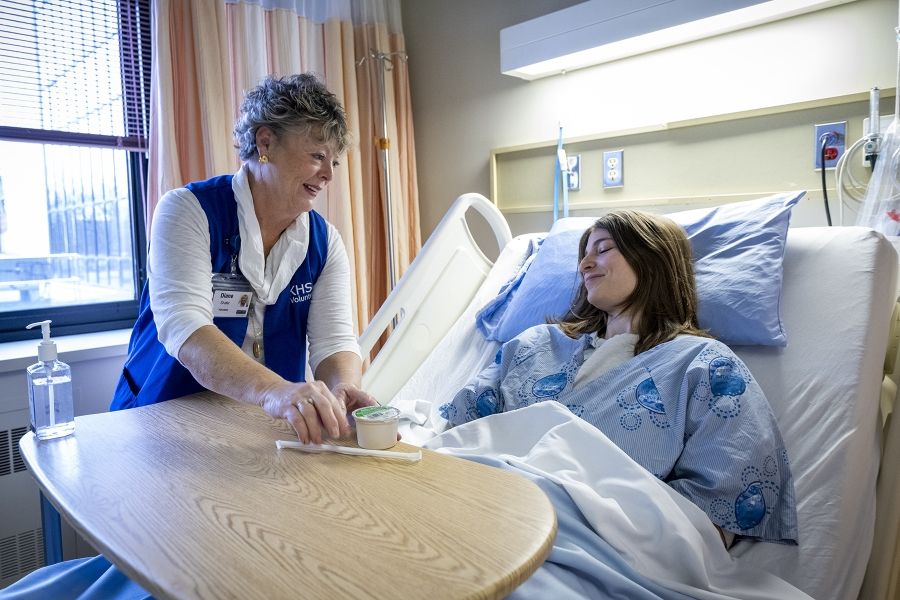 A volunteer is handing a patient, who is lying down in a bed, a snack.