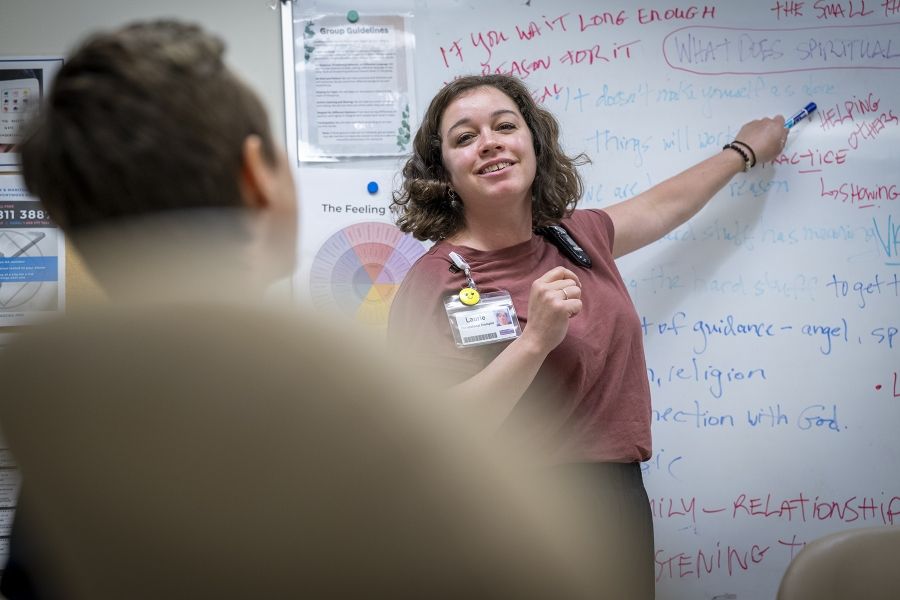 Laurie Hill is standing in front of a white board with a dry erase marker in her hand. She’s looking out at a colleague whose back is to the camera, and smiling while pointing at something on the board with the dry erase marker. She has short brown wavy hair and is wearing a dusty rose blouse and black pants.