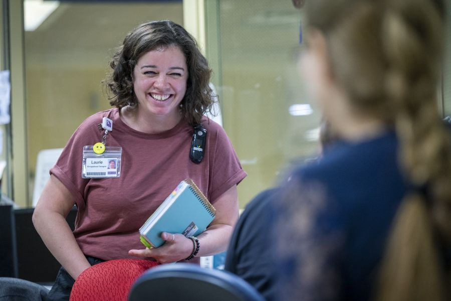 Laurie Hill is smiling and laughing while chatting with a colleague, whose back is facing the camera. Hill is holding her notebooks. She has short brown wavy hair and is wearing a dusty rose blouse and black pants.