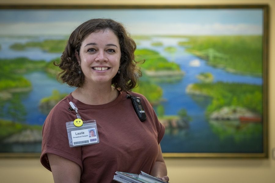 Laurie Hill is photographed in a hallway. She’s looking directly at the camera and smiling. She has short brown wavy hair and is wearing a dusty rose blouse and black pants.
