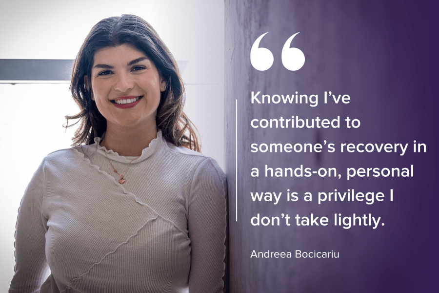 Andreea Bocicariu is standing against a wall and smiling directly into the camera. She has brown wavy hair, brown eyes and is wearing a white blouse with a gold heart necklace.