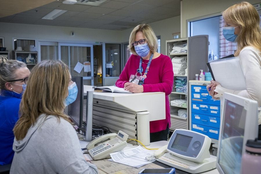 A wide shot of Clare Bowley who is standing at a care desk chatting with a few colleagues. Clare is taking notes in her black book. Clare has a blonde bob, blue eyes, has tortoise framed glasses on and a mask, and is wearing a pink sweater with her decorated KHSC lanyard on top.