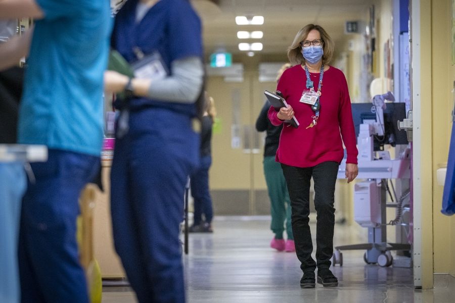 A wide shot of Clare Bowley walking down a unit hallway wearing a mask. Clare is holding her black book and calendar. Clare has a blonde bob, blue eyes, has tortoise framed glasses on, and is wearing a pink sweater with her decorated KHSC lanyard on top.