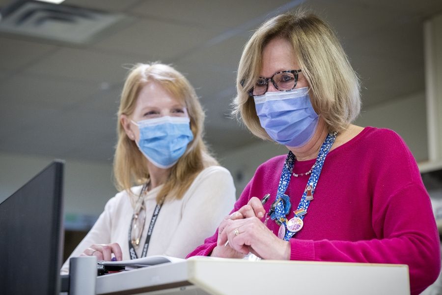 A wide shot of Clare Bowley and another social worker not in focus, standing and chatting at a care desk with masks one. Clare has a blonde bob, blue eyes, has tortoise framed glasses on, and is wearing a pink sweater with her decorated KHSC lanyard on top.