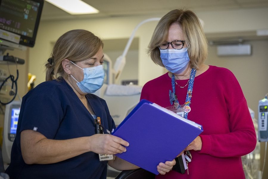 A wide shot of Clare Bowley going over a blue patient binder with the charge nurse in the Neonatal Intensive Care Unit. Clare has a blonde bob, blue eyes, has tortoise framed glasses on and a mask, and is wearing a pink sweater with her decorated KHSC lanyard on top. The charge nurse has blonde hair which is tied in a ponytail and is wearing a mask and navy blue scrubs.