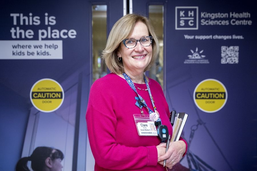 A wide shot of Clare Bowley standing outside the Pediatric unit and looking directly into the camera, smiling. Clare has a blonde bob, blue eyes, has tortoise framed glasses on, and is wearing a pink sweater with her decorated KHSC lanyard on top.
