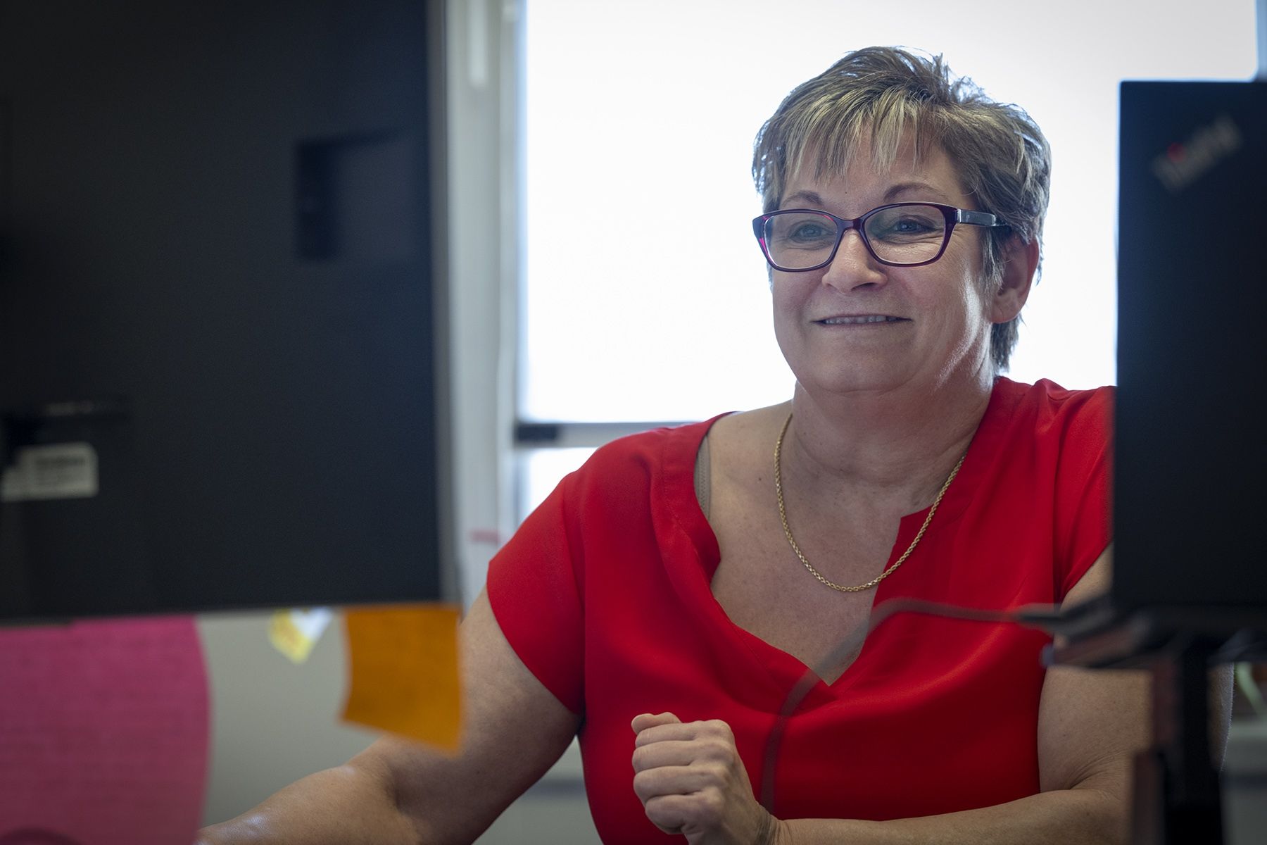 A shot of Marianne McKinney sitting at her desk in between two computer monitors. She’s smiling and looking at a computer monitor. McKinney has short dirty blonde hair, blue eyes, wears glasses and is dressed in a bright red blouse.