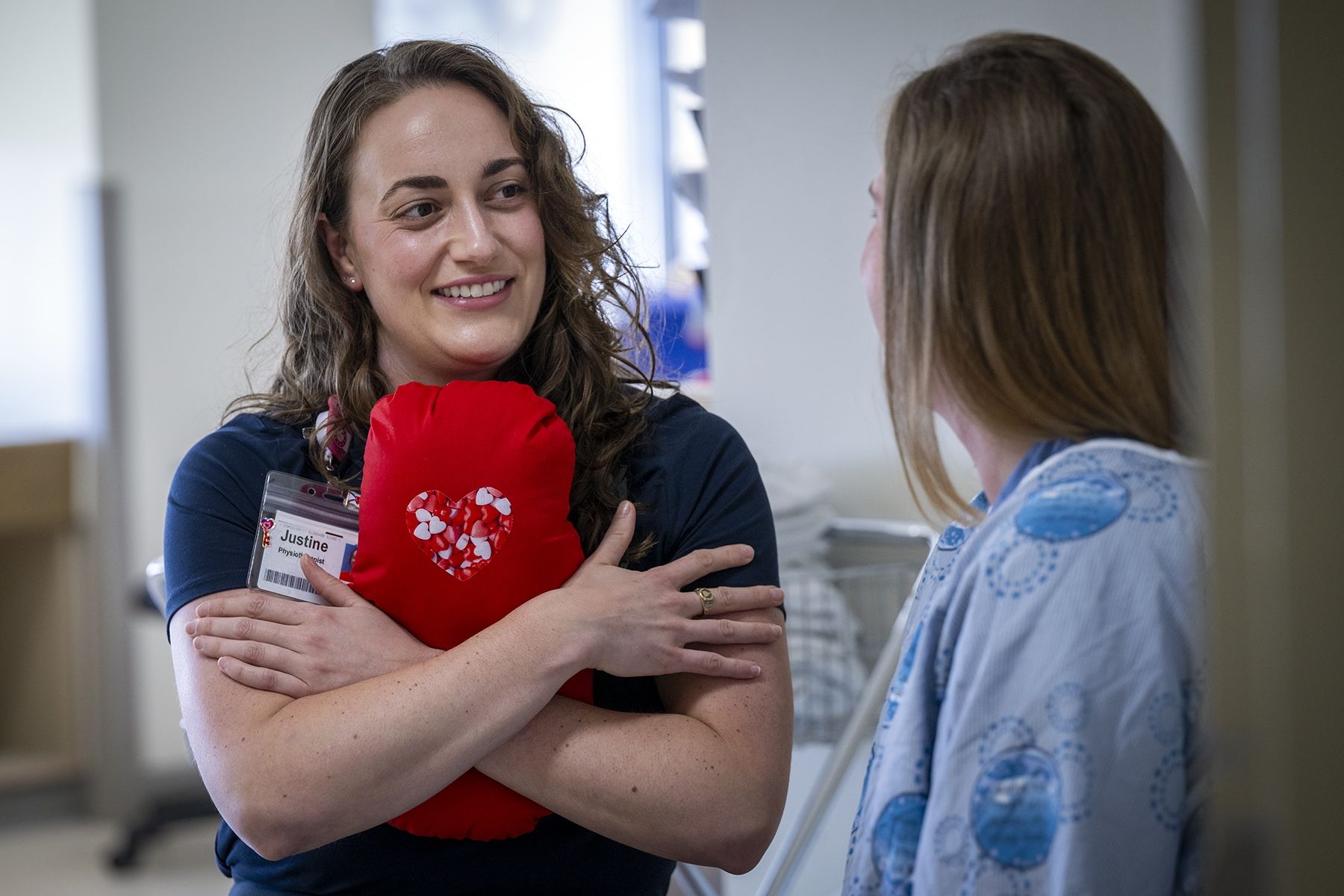 Justine White is standing, hugging a cardiac surgery pillow. She’s smiling and looking directly at another person whose back is to the camera. She has shoulder length, medium brown, wavy hair and brown eyes. She’s wearing a navy blue shirt with her KHSC ID badge.