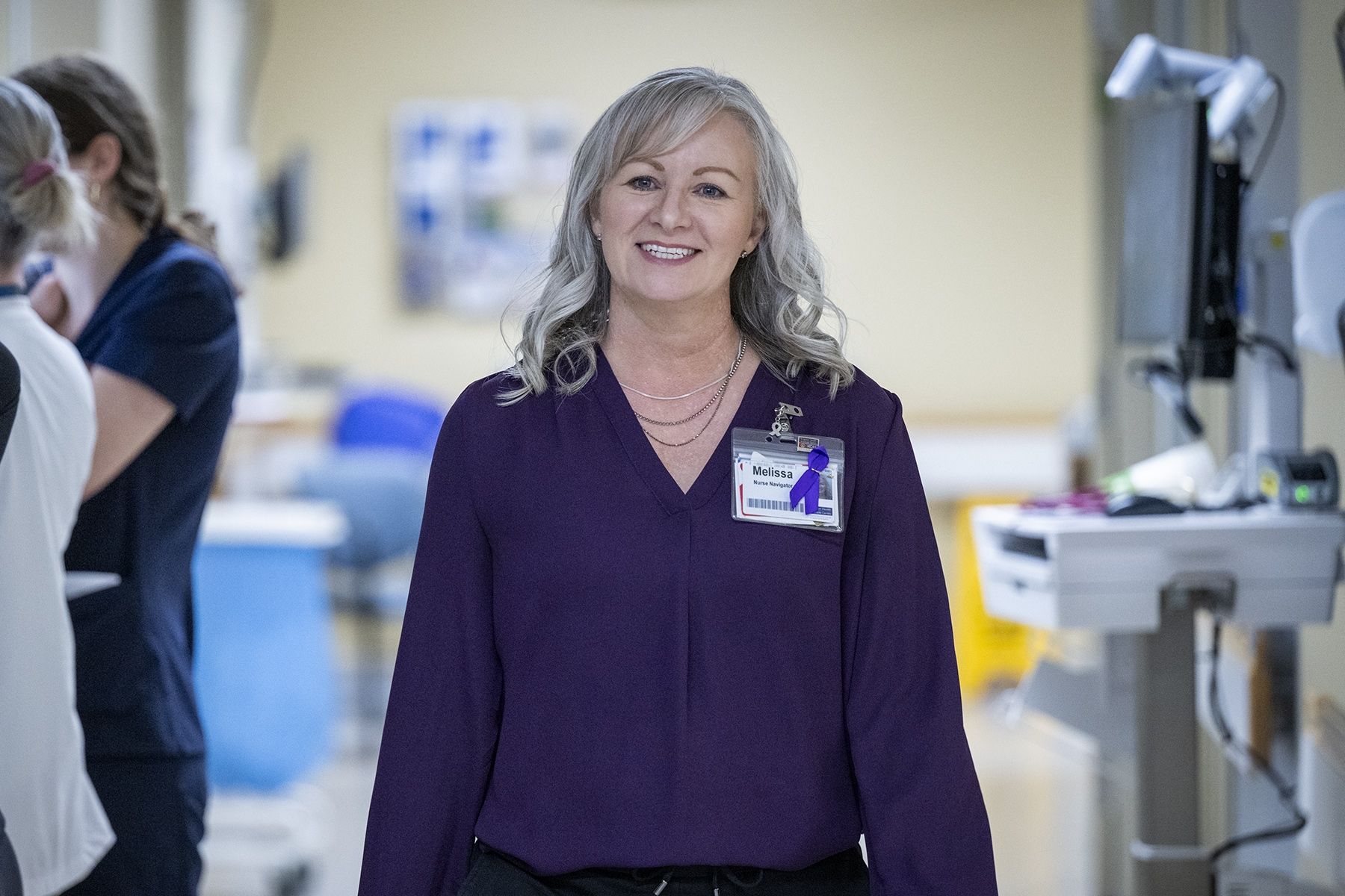 Melissa Meisner is walking down a hallway and smiling. Meisner has blue eyes, shoulder length, silver hair and is wearing a bright purple blouse.